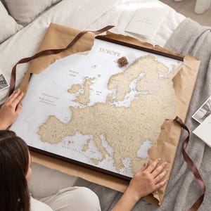 Può includere: Una mappa dell'Europa da grattare incorniciata con sfondo bianco e masse terrestri color oro. La mappa presenta il testo "EUROPE" e "OUR TRAVEL MAP" con una legenda per "Past Adventures" e "Adventures to Come" accanto a un piccolo contenitore di puntine.