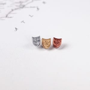 Puede incluir: Tres pequeños dijes de máscara de metal en colores plateado, dorado y cobre. Cada dije presenta un diseño de cara sonriente. Los dijes están dispuestos sobre una superficie blanca con un diseño de mapa tenue en el fondo.