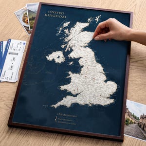 May include: A framed scratch-off map of the United Kingdom featuring a dark blue background and a white landmass. The map includes the text "United Kingdom" and "Our Adventures" with a legend for past and future travel pins.