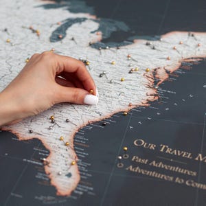 Può includere: Una mappa da parete degli Stati Uniti dal design scuro con terre grigio chiaro e oceani neri. La mappa presenta puntine metalliche color oro, argento e rame posizionate su vari stati. Il testo riporta Our Travel Map, Past Adventures.