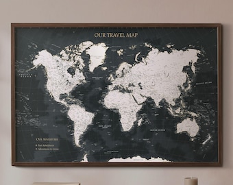May include: A rectangular world map framed in dark wood with a dark charcoal background and light-colored landmasses. The text OUR TRAVEL MAP is printed at the top center. The bottom left corner features the text OUR ADVENTURES with two bullet points.