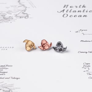 May include: Three small, metallic dog-shaped pins in gold, rose gold, and silver. The pins are detailed with visible features and are arranged on a white surface with a world map in the background. These pins are perfect for dog lovers.