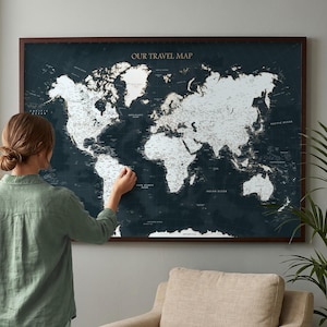 May include: A large rectangular wall map titled OUR TRAVEL MAP featuring a dark navy blue background with white landmasses and continents. The map displays geographical labels and borders in a clean font, framed in a dark brown wooden border.