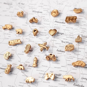 Puede incluir: Una colección de pequeños pines de metal en tono dorado con formas de diversos objetos, incluyendo vehículos, animales, un corazón, un mando de juego y una bicicleta, esparcidos sobre un mapa impreso del Medio Oeste de EE. UU. con nombres de ciudades.