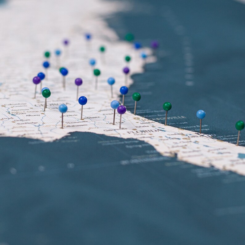 Puede incluir: Un mapa detallado de Florida y los estados circundantes, marcado con chinchetas de colores. Las chinchetas son de varios colores, incluyendo azul, verde y morado, indicando ubicaciones. El mapa es de un color neutro.