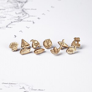 May include: A collection of small, gold-colored metal charms in various shapes, including a seashell, sailboat, turtle, compass, surfboard, lotus flower, palm tree, and fish. The charms are arranged on a white surface with a map design.