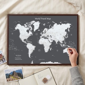 May include: A framed world travel map with a dark grey background and white continents. The text "World Travel Map" is at the top, and "Our Travels" with a legend is in the bottom left. Small pins mark locations across the landmasses.