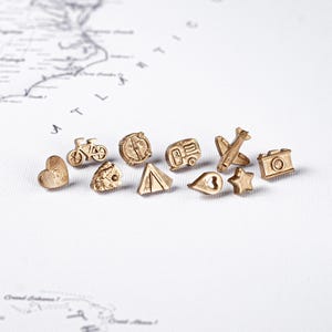 Può includere: Una collezione di piccoli ciondoli dorati a tema viaggio. I ciondoli includono un cuore, una bicicletta, una bussola, un rimorchio, un aereo, una fotocamera, una tenda e una stella. I ciondoli sono disposti su una superficie bianca con una mappa sullo sfondo.