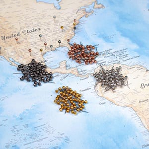 Op de afbeelding: Een wereldkaart met beige landmassa's en blauwe oceanen, met diverse losse punaises in de Verenigde Staten en vier afzonderlijke groepen metalen punaises in koper, zwart, zilver en goud die op de kaart zijn gerangschikt.