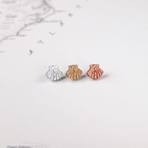 May include: Three small seashell-shaped charms in silver, gold, and rose gold. The charms are arranged in a row against a white background with a faint map design. These decorative charms are ideal for jewellery making or craft projects.