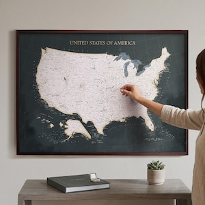 May include: A framed wall map of the United States of America featuring a dark charcoal background with a light-coloured map overlay. The map displays state borders, major cities, and geographical features, enclosed in a dark brown wooden frame.