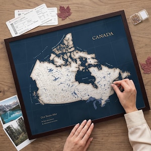 Puede incluir: Un mapa de pared enmarcado de Canadá con fondo azul oscuro y masas terrestres de color crema que presenta el texto CANADA y Our Travel Map. El mapa incluye una leyenda para aventuras pasadas y futuros viajes, junto a dos tarjetas de embarque.