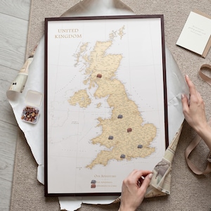 Puede incluir: Un mapa enmarcado del Reino Unido con fondo crema y masas terrestres doradas. El mapa presenta el texto "United Kingdom" y "Our Adventures" con una leyenda que muestra dos pines de colores. Varios pines están colocados sobre el mapa.