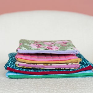 May include: A stack of five quilted fabric pouches in various colors and patterns. The top pouch has a floral print with pink and green hues, while others feature stripes and geometric designs. Each pouch has a zipper closure.