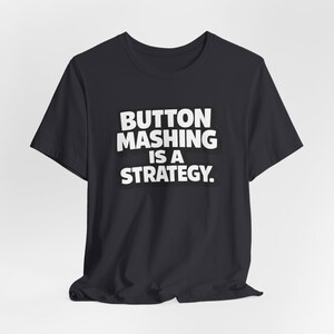May include: Black t-shirt with the white text "BUTTON MASHING IS A STRATEGY." The text is in a bold, sans-serif font with a black outline. The shirt is made of a soft material and has short sleeves.