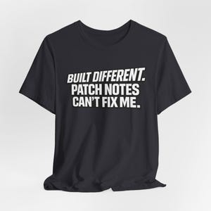 May include: Black t-shirt featuring the white text "BUILT DIFFERENT. PATCH NOTES CAN'T FIX ME." in a bold, sans-serif font. The shirt has a crew neck and short sleeves.