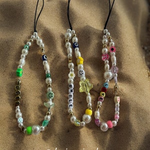 May include: Three beaded phone charms with white pearls, gold beads, and colorful accents. One charm spells "LUCKY", another "SMILE", and the third "LOVE". Each charm has a black cord loop for attachment. The charms are displayed on a sandy surface.