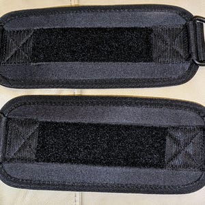 May include: Two black ankle straps with D-rings. Each strap has a rectangular design with a central Velcro strip for secure fastening. The straps are made of a durable, textured material, suitable for fitness activities.
