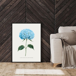 Puede incluir: Un cuadro enmarcado con una ilustración detallada de una flor de hortensia azul. La flor tiene una gran floración esférica y hojas verdes. La impresión está sobre un fondo blanco, con texto en la parte inferior. El marco es negro.