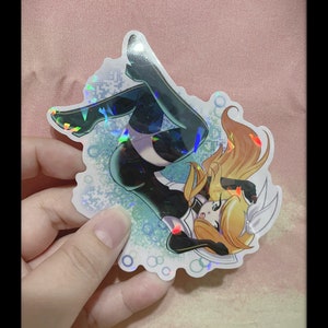 Może przedstawiać: Holograficzna naklejka przedstawiająca postać w stylu anime w dynamicznej pozie. Postać ma na sobie czarno-biały strój z blond włosami. Naklejka ma błyszczący, opalizujący efekt.