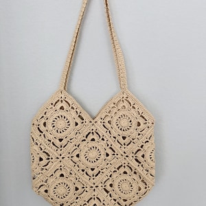 May include: A beige crochet tote bag with a geometric floral pattern. The bag has two long handles and is hanging against a light grey wall. The bag's design features repeating circular and square motifs.
