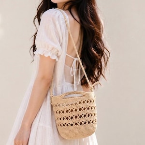 May include: A beige, crocheted shoulder bag with a handle and a strap. The bag has an open weave design, and is hanging from the shoulder of a person wearing a white dress.