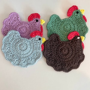 May include: Four crocheted chicken-shaped coasters in different colors: purple, green, blue, and brown. The coasters are made with a circular design and have a red comb and yellow beak.