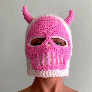 Può includere: Un passamontagna all'uncinetto fatto a mano in bianco e rosa, con un design a teschio con corna. La maschera ha aperture per occhi e bocca, e i dettagli del teschio sono di un rosa vibrante.