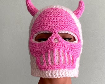 May include: A handmade crochet balaclava in white and pink, featuring a skull design with horns. The mask has eye and mouth openings, and the skull details are in a vibrant pink color.