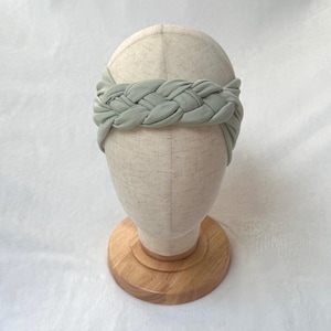 May include: A sage green fabric headband featuring a thick, braided design that wraps around the head. The headband is displayed on a neutral-colored mannequin head stand with a circular wooden base, showcasing the soft texture of the material.