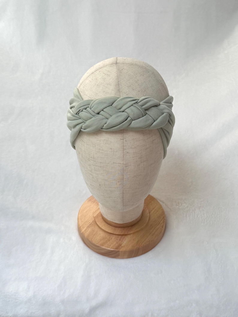 May include: A sage green fabric headband featuring a thick, braided design that wraps around the head. The headband is displayed on a neutral-colored mannequin head stand with a circular wooden base, showcasing the soft texture of the material.