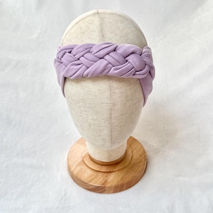 May include: A wide, light purple fabric headband featuring a thick, braided design across the front. The headband is displayed on a neutral-colored mannequin head stand with a circular wooden base, showcasing the soft texture of the material.