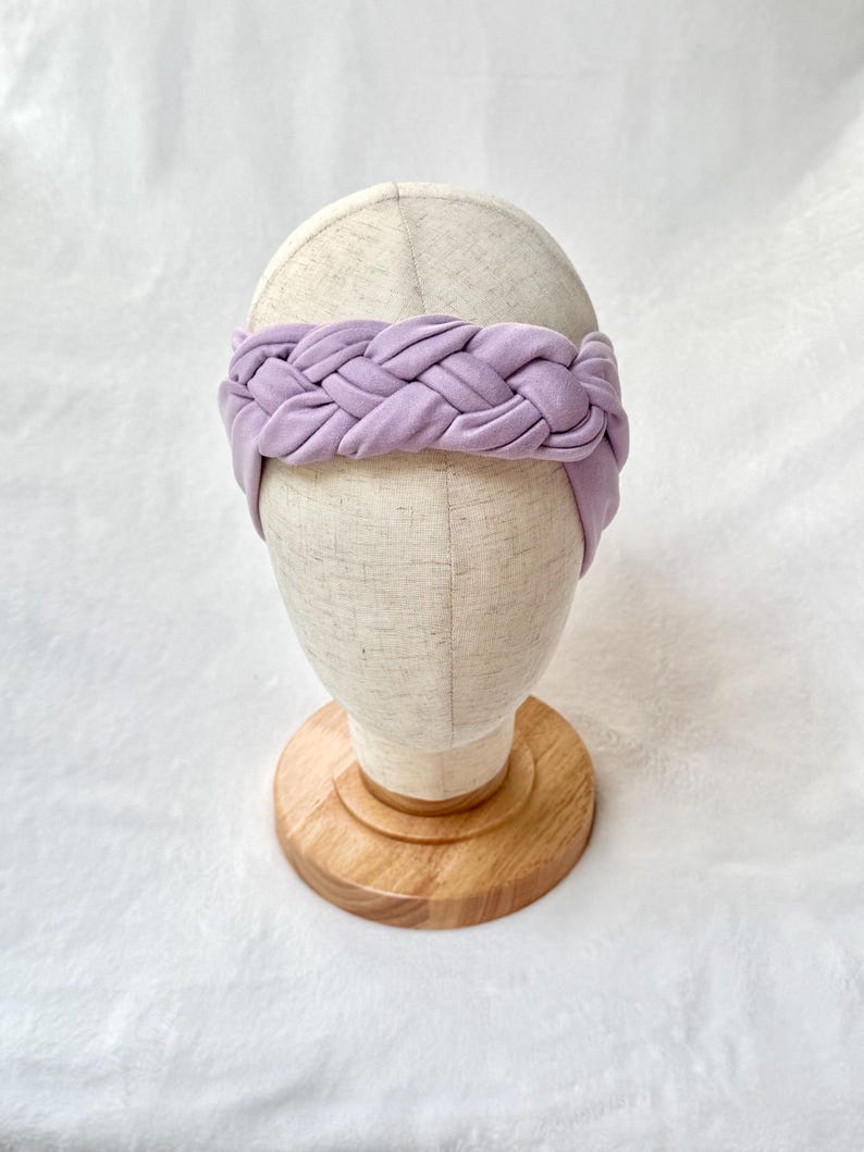 May include: A wide, light purple fabric headband featuring a thick, braided design across the front. The headband is displayed on a neutral-colored mannequin head stand with a circular wooden base, showcasing the soft texture of the material.