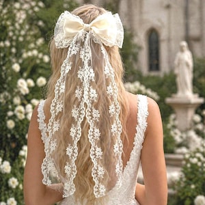 Könnte beinhalten: Cremefarbener Braut-Haarschmuck mit einer großen Satinschleife und einem zarten Spitzenschleier. Der Schleier ist mit floralen Mustern verziert und fällt über den Rücken. Der Haarschmuck ist für eine Hochzeit oder einen besonderen Anlass gedacht.
