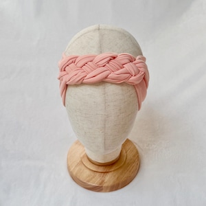 May include: A soft, light pink fabric headband featuring a wide, braided design. The headband is displayed on a neutral-toned mannequin head stand with a circular wooden base, showcasing the smooth texture and uniform color of the material.