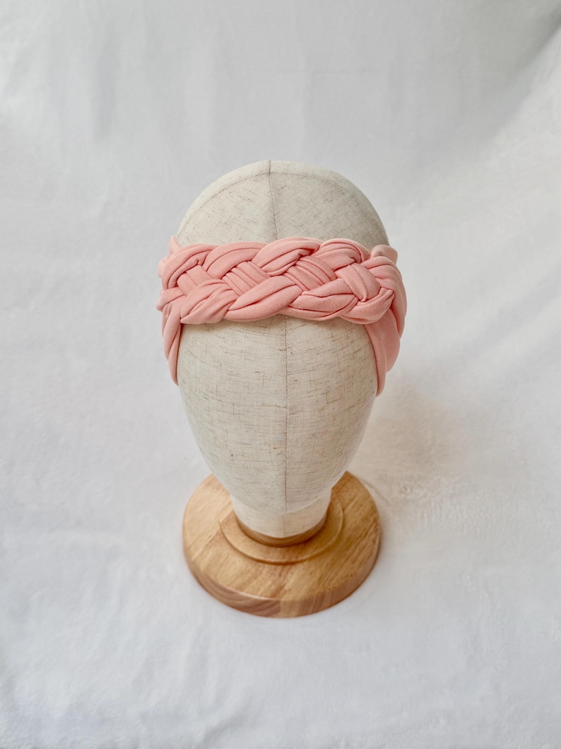 May include: A soft, light pink fabric headband featuring a wide, braided design. The headband is displayed on a neutral-toned mannequin head stand with a circular wooden base, showcasing the smooth texture and uniform color of the material.