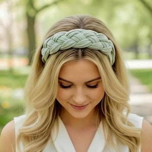 May include: A sage green headband featuring a thick, braided fabric design. The headband is worn across the top of the head, showcasing a wide, woven texture with a soft, matte finish that sits securely against the hair in a uniform pattern.