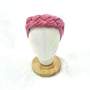 May include: A dusty rose pink headband featuring a thick, braided fabric design. The headband is displayed on a neutral-colored mannequin head stand, showcasing the intricate woven texture and the soft, matte finish of the fabric material.