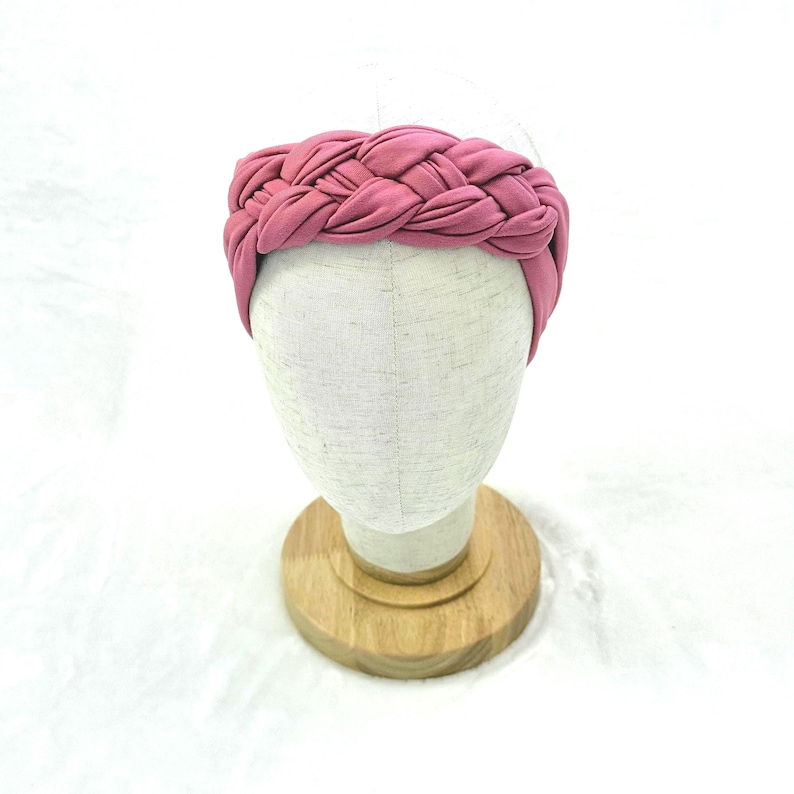 May include: A dusty rose pink headband featuring a thick, braided fabric design. The headband is displayed on a neutral-colored mannequin head stand, showcasing the intricate woven texture and the soft, matte finish of the fabric material.