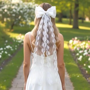 Könnte beinhalten: Ein weißes Haaraccessoire mit einer großen Satinschleife, die an einem langen, transparenten Spitzenschleier befestigt ist. Der Schleier ist mit aufwendigen Blumenstickereien an den Rändern verziert und fällt über den Rücken.