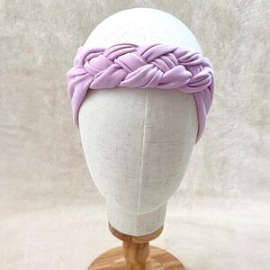 May include: A light lavender headband featuring a thick, braided fabric design. The headband is constructed from a soft, matte material and wraps around the head with a wide, uniform band that showcases a prominent, central woven braid detail.