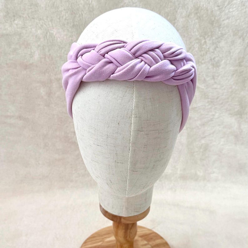 May include: A light lavender headband featuring a thick, braided fabric design. The headband is constructed from a soft, matte material and wraps around the head with a wide, uniform band that showcases a prominent, central woven braid detail.