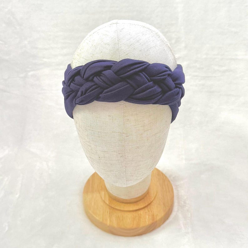 May include: A dark navy blue headband featuring a thick, braided fabric design. The headband is displayed on a neutral-colored mannequin head stand, showcasing the intricate woven texture and the solid, deep blue color of the soft fabric material.