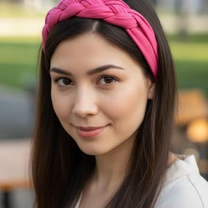 May include: A bright pink fabric headband featuring a thick, braided design. The headband is constructed from a smooth, solid-colored material that sits across the top of the head, showcasing a woven, multi-strand plait pattern throughout.