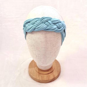 May include: A light blue headband featuring a thick, braided fabric design. The headband is displayed on a neutral-colored mannequin head stand, showcasing the soft, matte texture of the material and the intricate, interwoven pattern of the braid.
