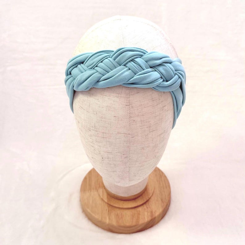 May include: A light blue headband featuring a thick, braided fabric design. The headband is displayed on a neutral-colored mannequin head stand, showcasing the soft, matte texture of the material and the intricate, interwoven pattern of the braid.