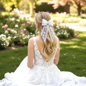 May include: A bride in a white lace wedding dress with a low back and a large white lace bow in her hair. The bow features a pearl centre and long, flowing lace tails. The bride is seated on green grass in a garden setting.