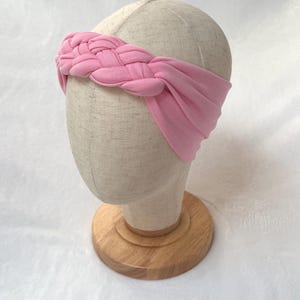 May include: A light pink fabric headband featuring a braided design across the front. The headband is made of a soft, solid-colored material that wraps around the head, with a wide, smooth band extending from the central braid to the sides.