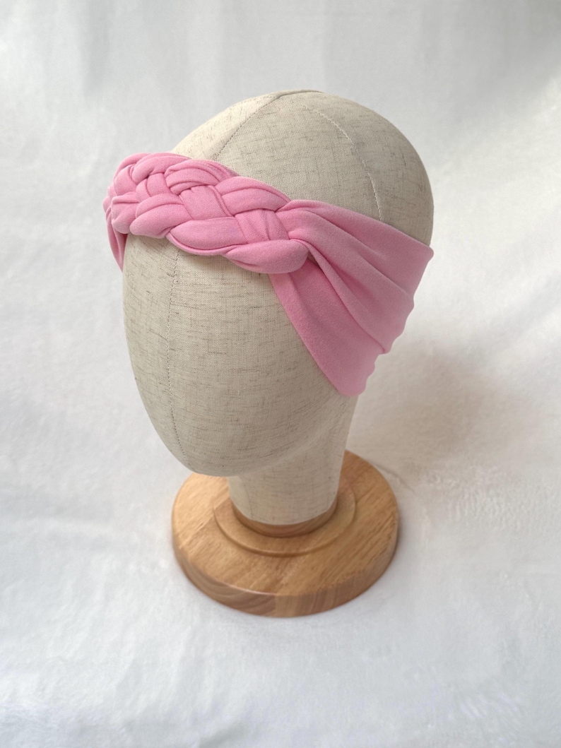 May include: A light pink fabric headband featuring a braided design across the front. The headband is made of a soft, solid-colored material that wraps around the head, with a wide, smooth band extending from the central braid to the sides.