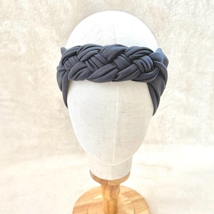 May include: A dark gray fabric headband featuring a thick, braided design that wraps around the crown. The headband is displayed on a plain, off-white mannequin head, showcasing the uniform color and the intricate, woven texture of the material.